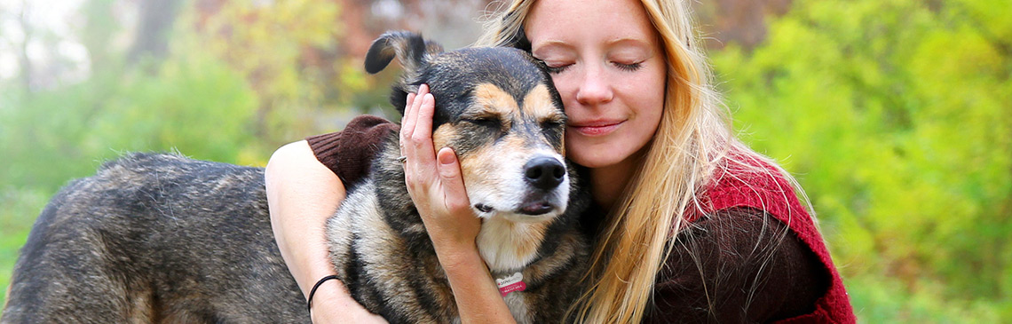 A happy, peaceful young woman has stopped walking her senior dog along a path in the autumn forest to hug him lovingly.